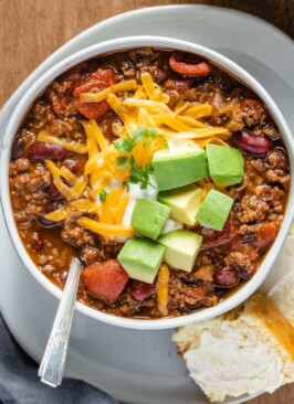 Venison chili, above shot, in a bowl topped with shredded cheddar cheese, chopped avocado and a dollop of sour cream