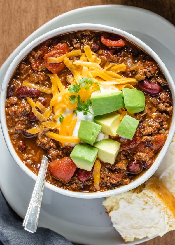 Venison chili, above shot, in a bowl topped with shredded cheddar cheese, chopped avocado and a dollop of sour cream