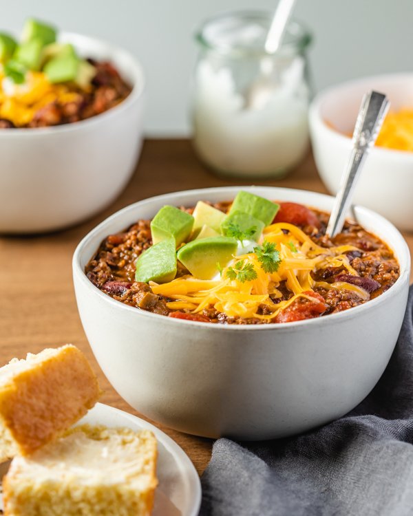 Bowl of venison chili with spoon in bowl topped with shredded cheddar cheese, avocado and a side of corn bread