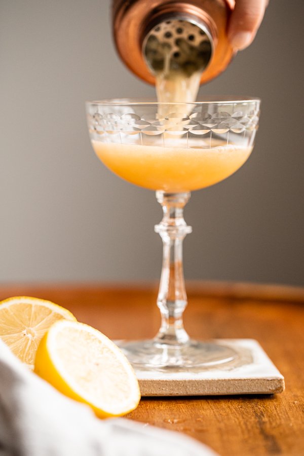 a cocktail shaker pouring a whiskey sour into a coupe glass