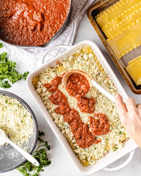 scooping red sauce over cheese in a lasagna pan