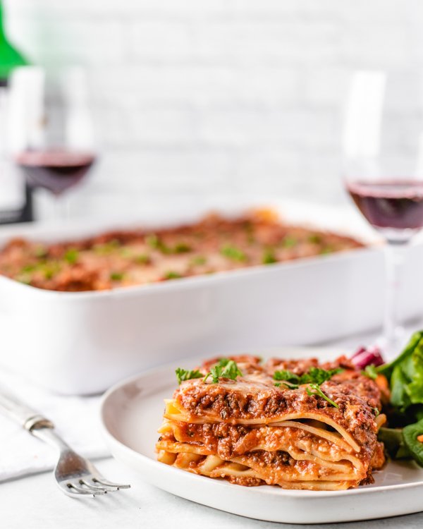 a slice of venison lasagna on a white plate with a dish and wine in the background