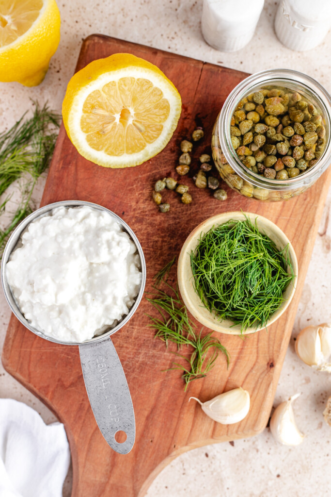 Cottage Cheese in measuring cup, Fresh Dill, Garlic, half a lemon, and Capers in a jar on a wooden cutting board