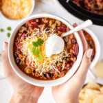 Hands holding a white bowl of Slow Cooker Venison Chili topped with cheese, sour cream and cilantro