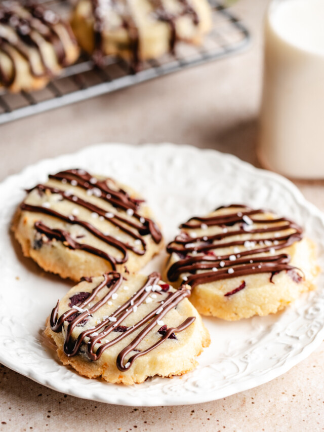 Three baked Shortbread Cookies with Orange & Cranberry on a white plate with milk and cooling rack in background