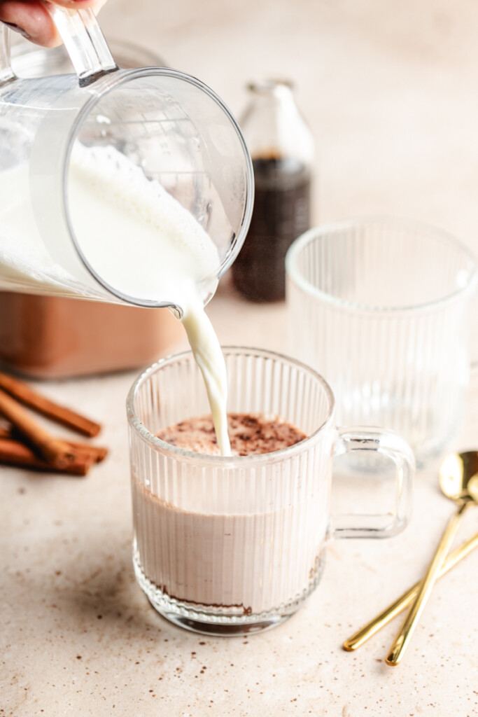 milk being poured over hot cocoa mix in glass mug