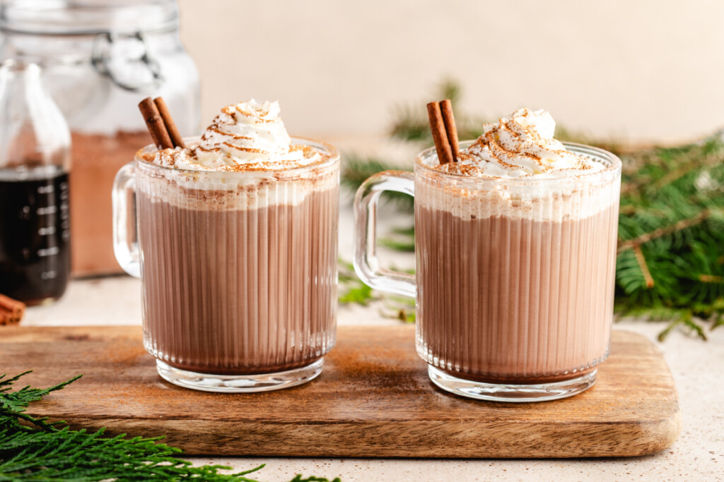 healthier hot chocolate mix in a glass mug on a brown cutting board topped with a cinnamon stick, whipped cream and cinnamon. Vanilla extract and hot cocoa mix in background.