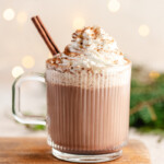 healthier hot chocolate mix in a glass mug topped with whipped cream, cinnamon and a cinnamon stick with lights in the background