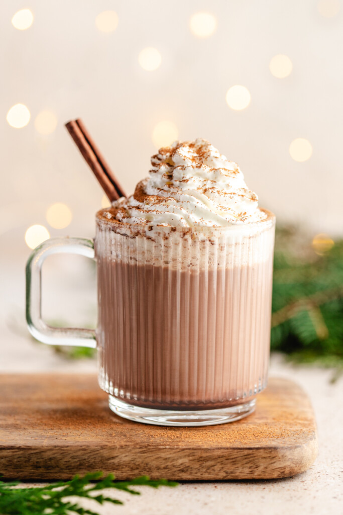 healthier hot chocolate mix in a glass mug topped with whipped cream, cinnamon and a cinnamon stick with lights in the background