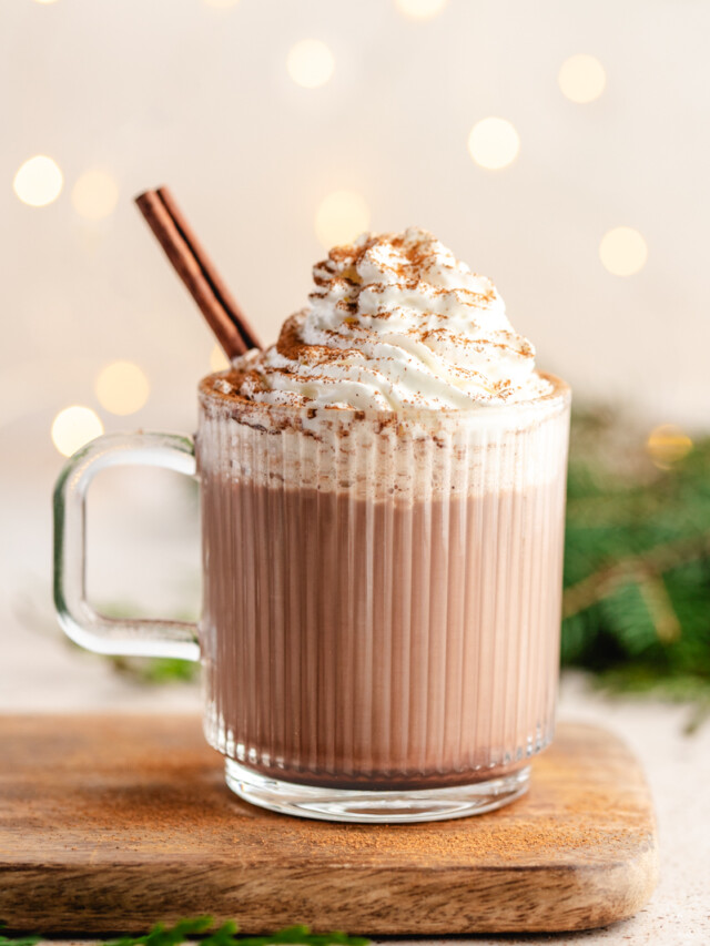 healthier hot chocolate mix in a glass mug topped with whipped cream, cinnamon and a cinnamon stick with lights in the background