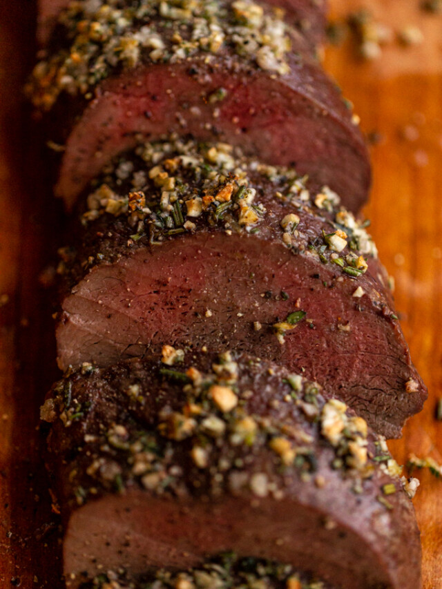 roasted venison backstrap recipe sliced on a wood board with herbs and garlic