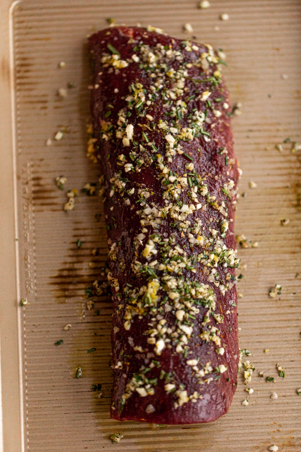 venison loin covered in garlic and herbs on a sheet pan