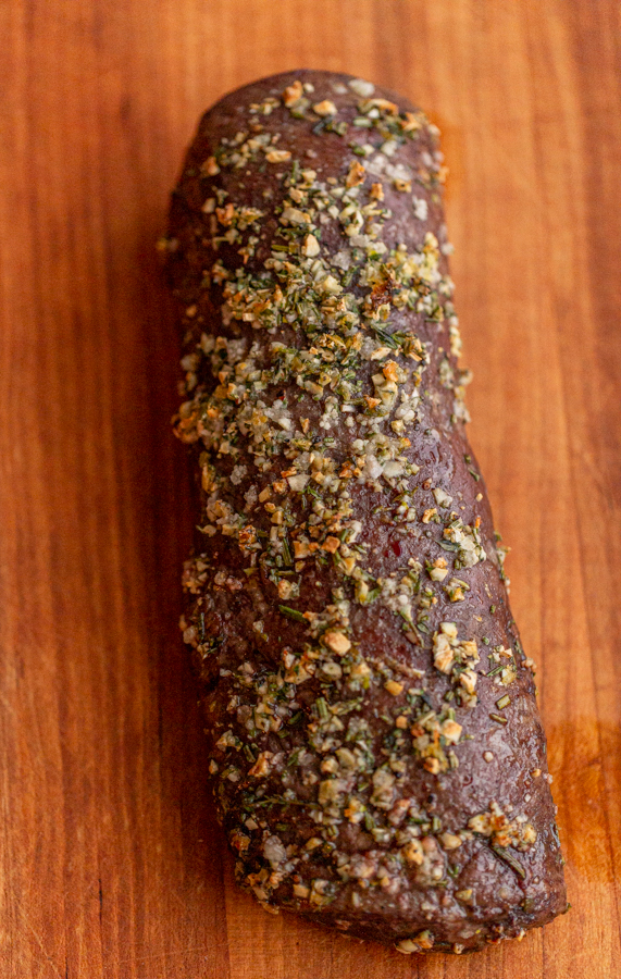 roasted venison backstrap recipe on a wood board with herbs and garlic