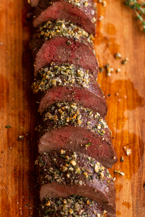 roasted venison backstrap recipe sliced on a wood board with herbs and garlic