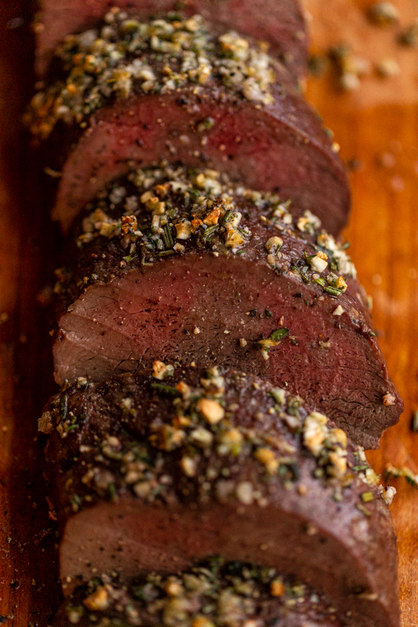 roasted venison backstrap recipe sliced on a wood board with herbs and garlic