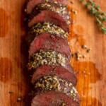 roasted venison backstrap recipe sliced on a wood board with herbs and garlic