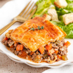 Venison Pot Pie with golden and flaky baked puff pastry topping served on a white plate with a green salad in the background