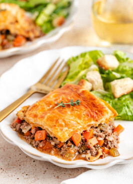 Venison Pot Pie with golden and flaky baked puff pastry topping served on a white plate with a green salad and white wine in the background