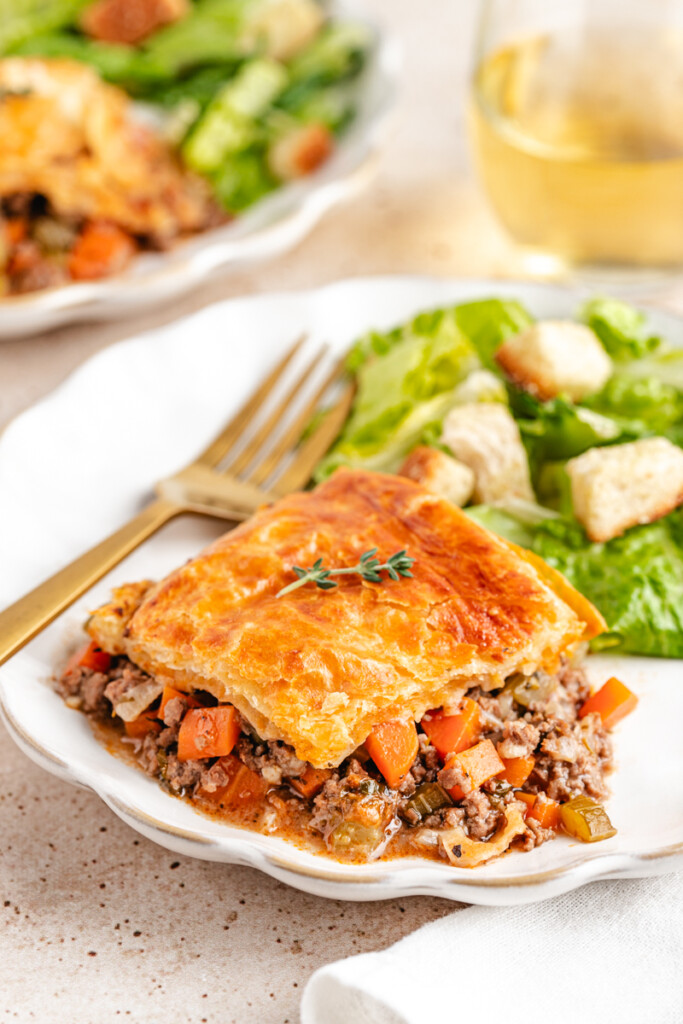 Venison Pot Pie with golden and flaky baked puff pastry topping served on a white plate with a green salad and white wine in the background