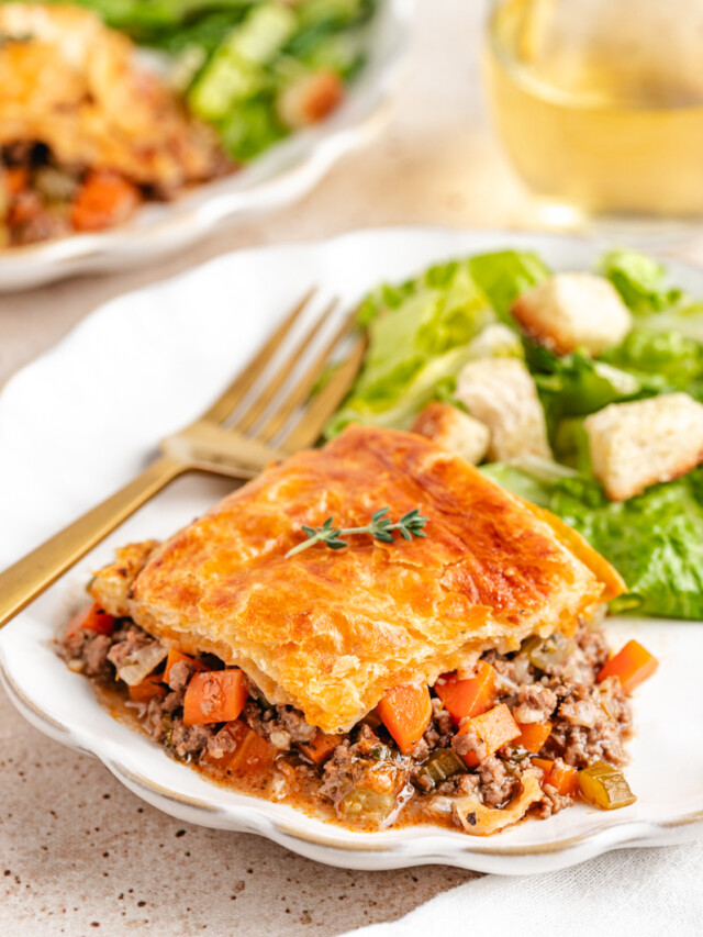 Venison Pot Pie with golden and flaky baked puff pastry topping served on a white plate with a green salad and white wine in the background
