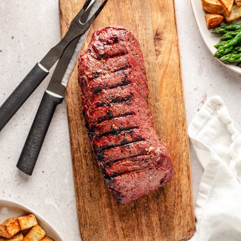 A grilled backstrap on a wooden cutting board with a carving knife and fork