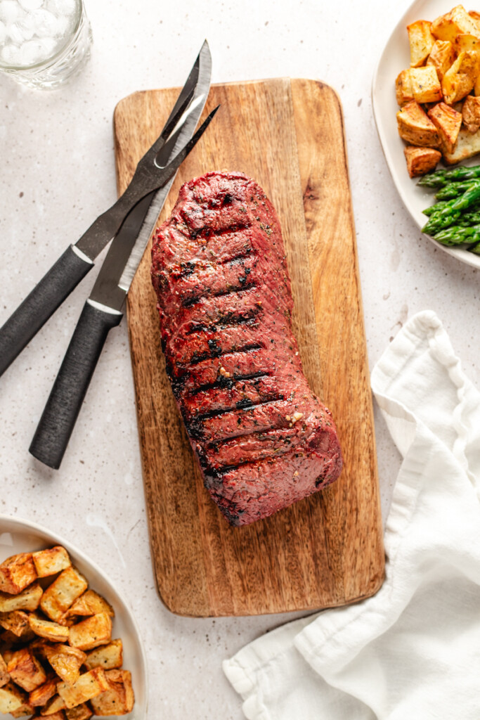 A grilled backstrap on a wooden cutting board with a carving knife and fork and plates surrounding it with potatoes and asparagus on them