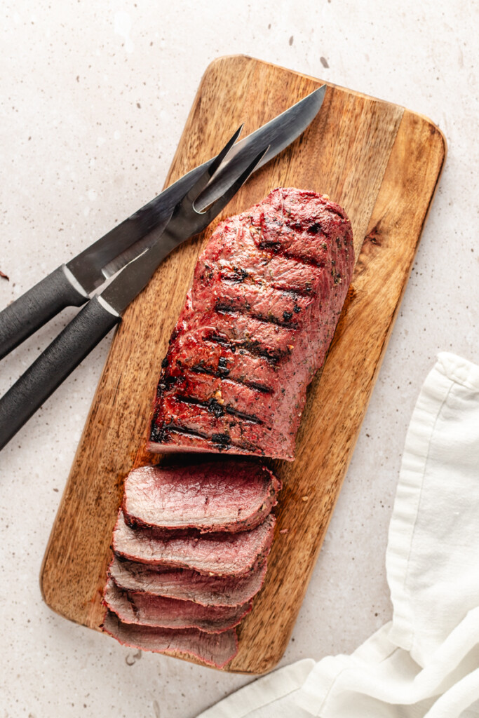 Sliced grilled venison backstrap on wooden cutting board with knife and carving fork