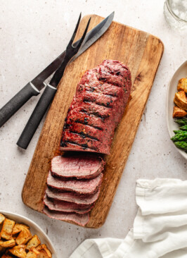 Sliced grilled venison backstrap on wooden cutting board with plates around it