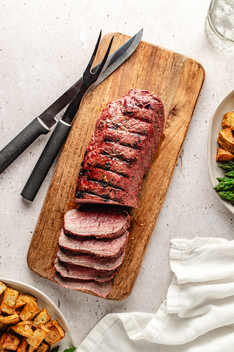 Sliced grilled venison backstrap on wooden cutting board with plates around it