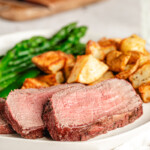 Slices of medium rare steak on a white plate with sides of asparagus and potatoes