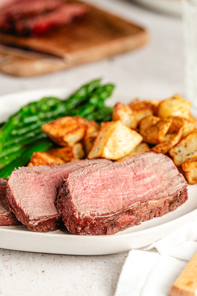 Slices of medium rare steak on a white plate with sides of asparagus and potatoes