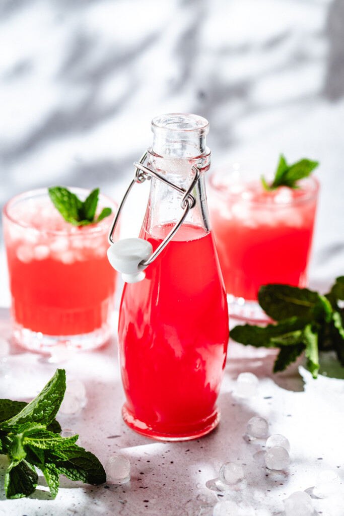 Strawberry Rhubarb Syrup in a glass flip top jar with two drinks in glasses behind it