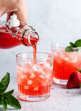 Strawberry Rhubarb Syrup being poured over sparkling water in a glass with pebble ice