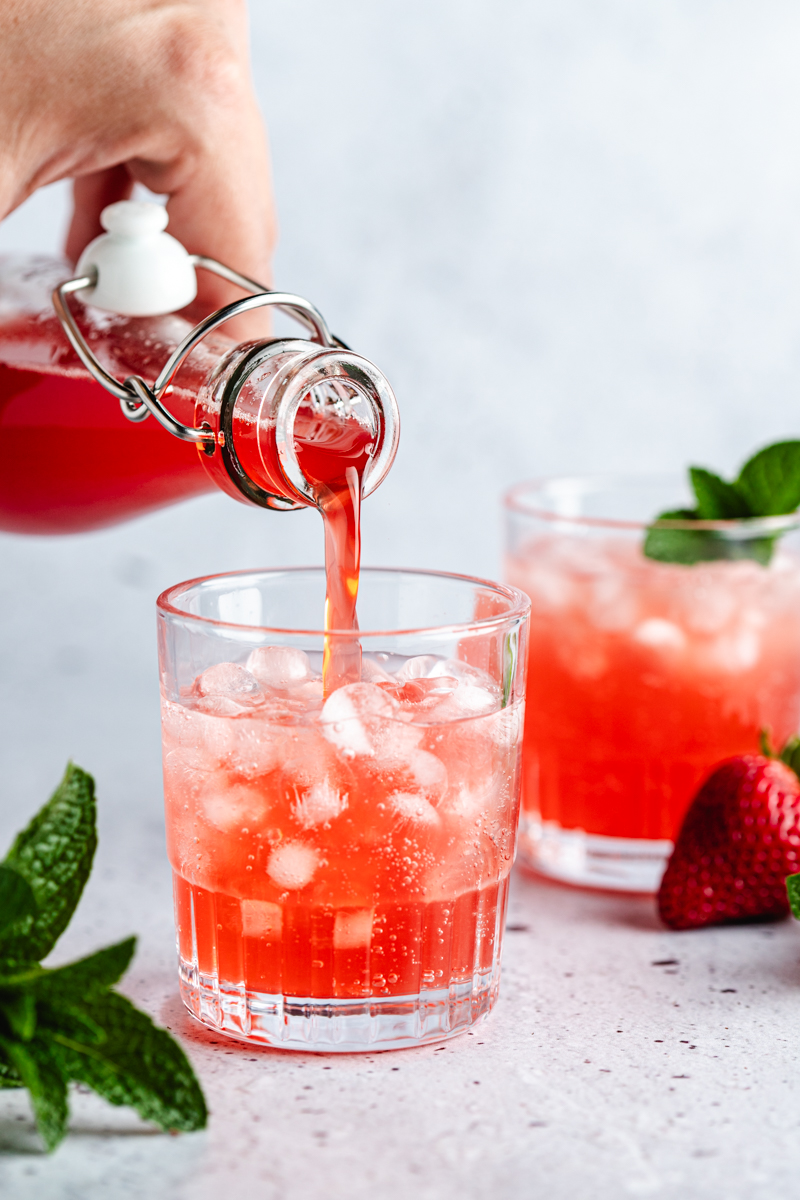Strawberry Rhubarb Syrup being poured over sparkling water in a glass with pebble ice