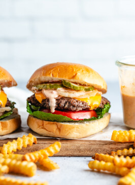 Venison smash burger on a wooden board surrounded by French fries and burger sauce