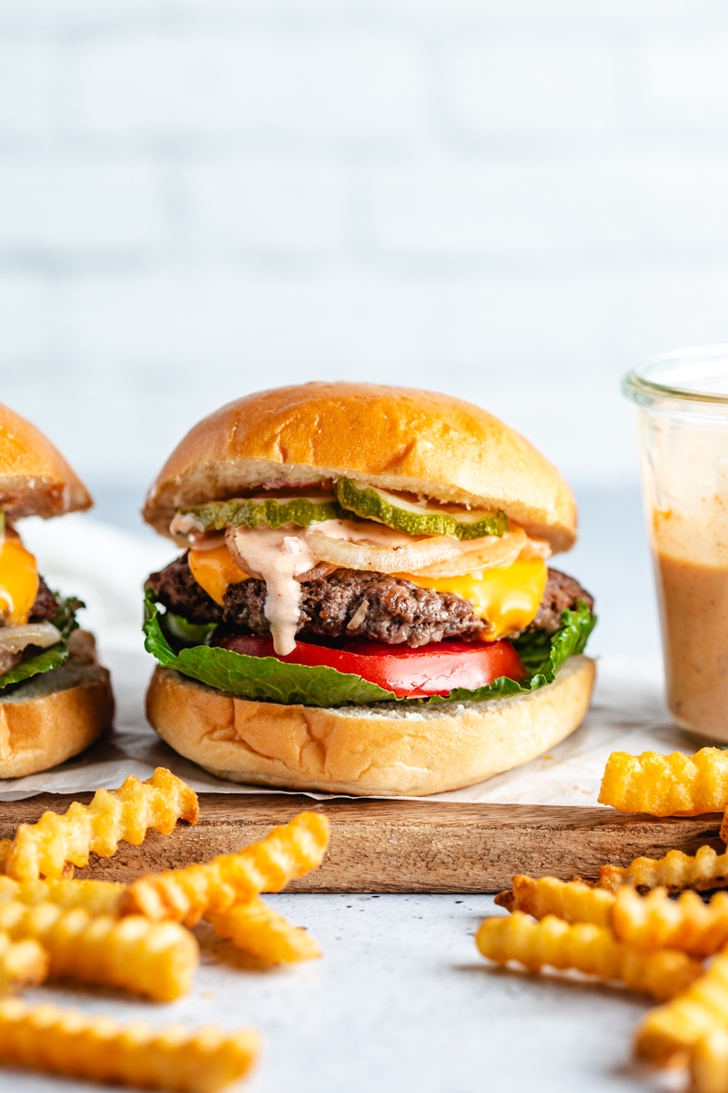 Venison smash burger on a wooden board surrounded by French fries and burger sauce