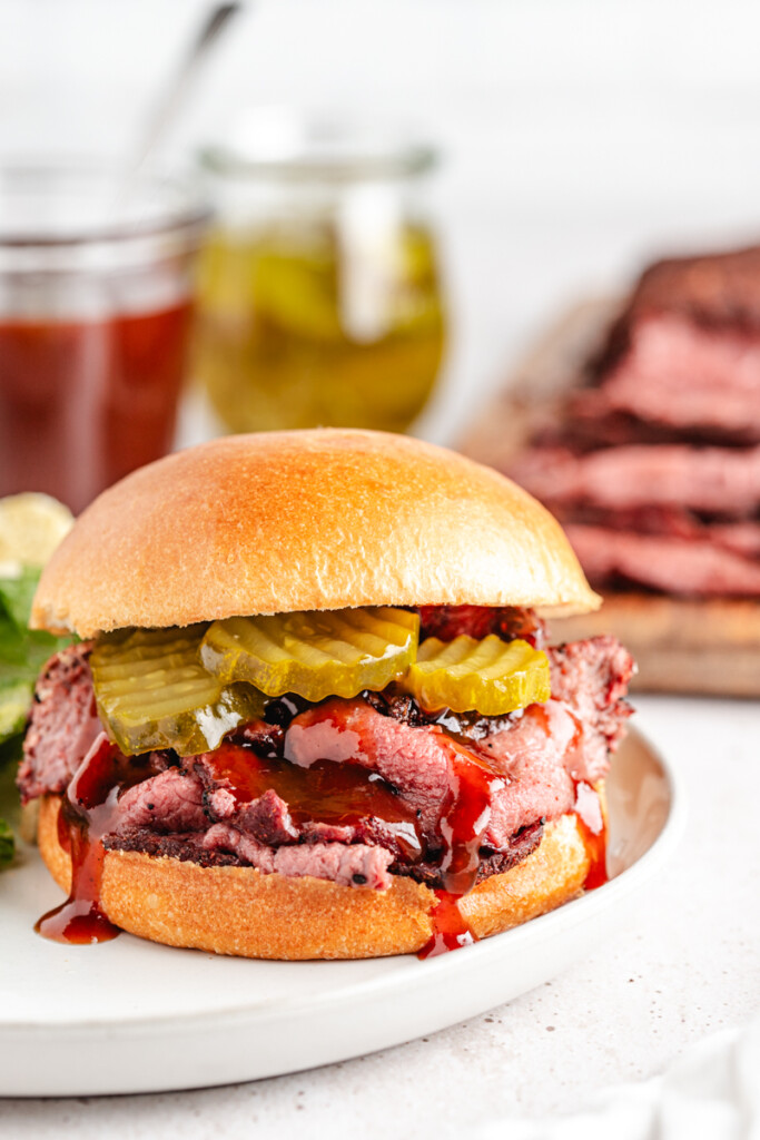 A sandwich with thinly sliced smoked venison, bbq sauce and pickles on a white plate