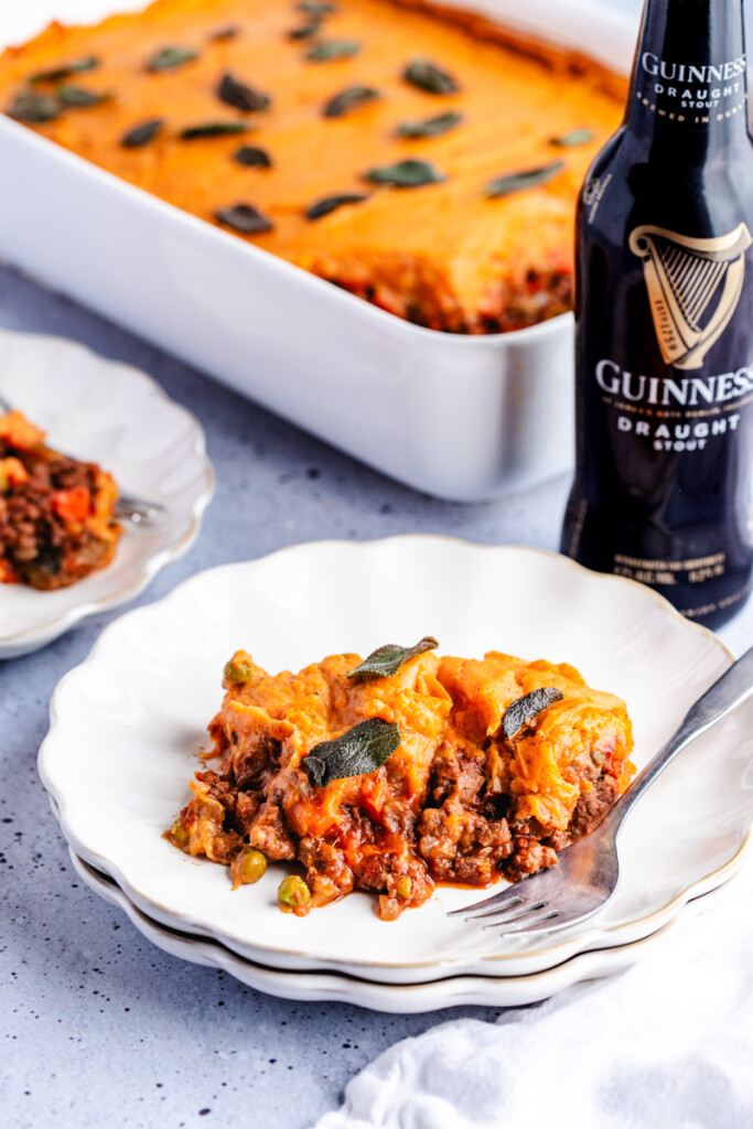 A serving of Sweet Potato Shepherd's Pie on a white plate garnished with crispy sage next to a stout beer