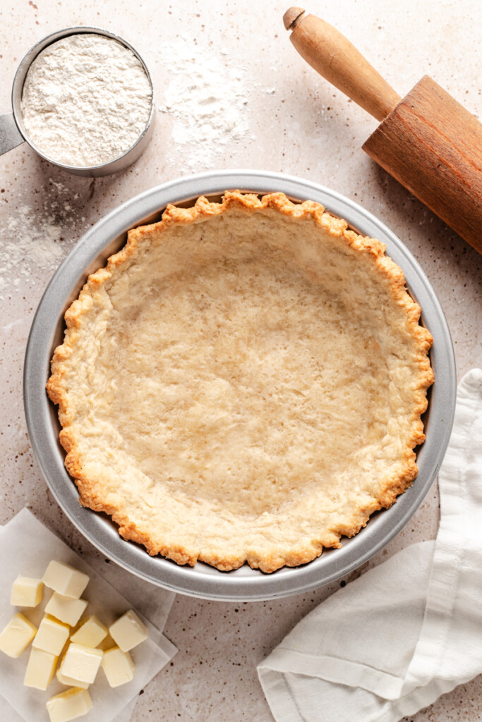 Homemade pie crust baked in a pie pan with a rolling pin, flour and cubed butter surrounding it