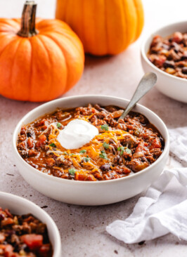 A white bowl of pumpkin chili topped with sour cream, shredded cheese and cilantro with orange pumpkins in the background