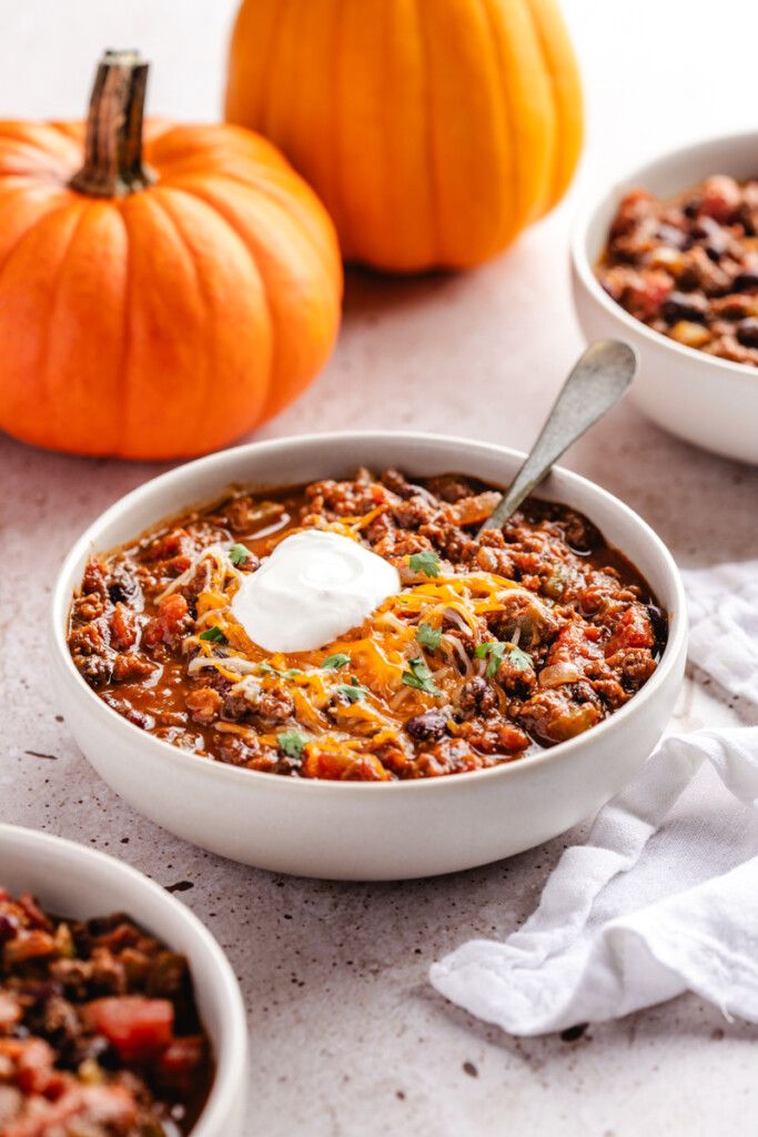 A white bowl of pumpkin chili topped with sour cream, shredded cheese and cilantro with orange pumpkins in the background