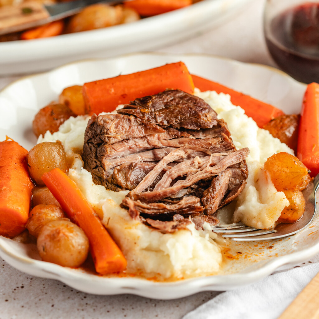 Close up shot of Beer Braised Venison Roast with pearl onions and carrots served over mashed potatoes on a white plate
