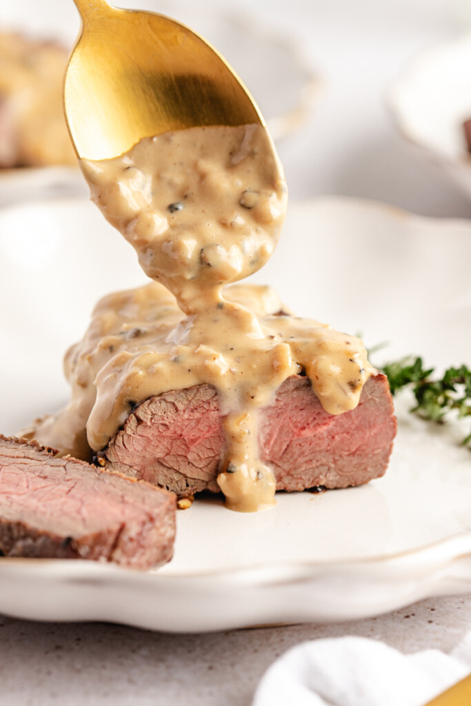 A gold spoon adding sauce to a Venison Au Poivre steak on a white plate
