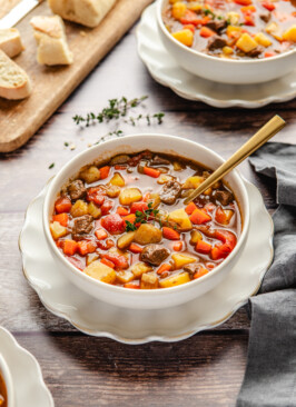 Venison stew in a white bowl surrounded by crusty bread and thyme