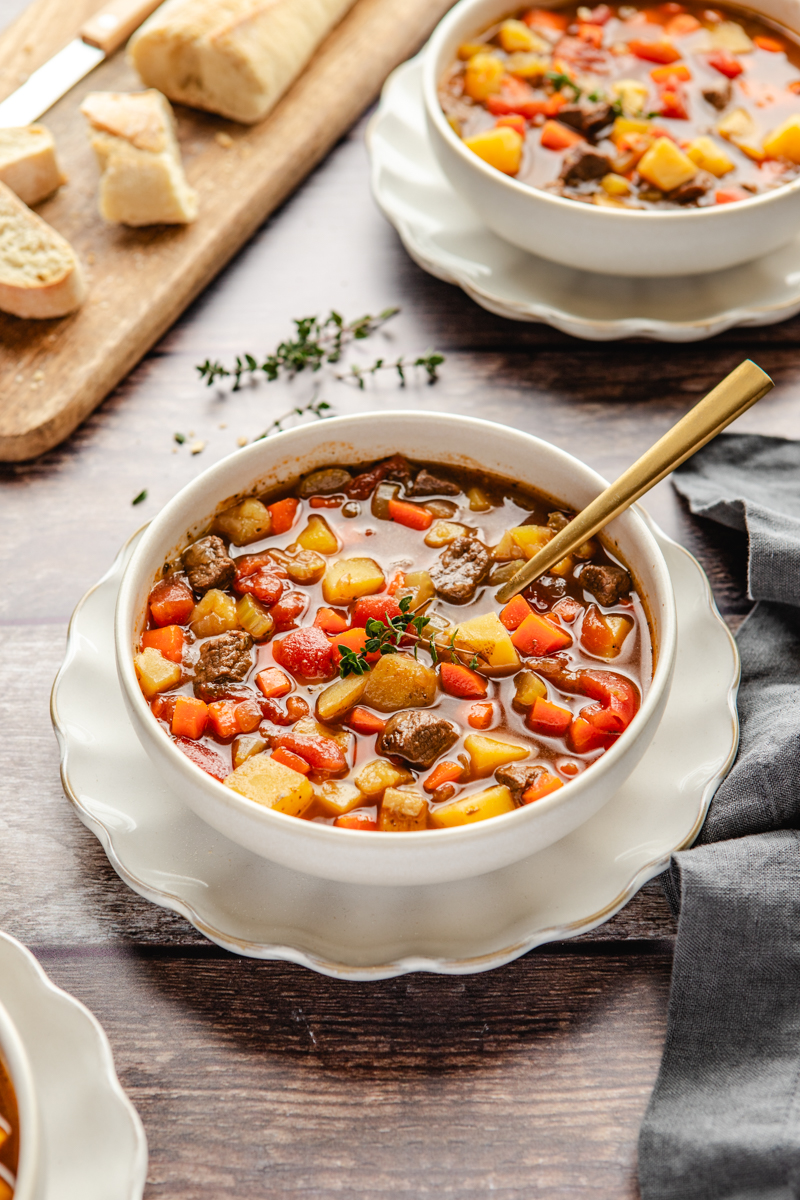 Venison stew in a white bowl surrounded by crusty bread and thyme
