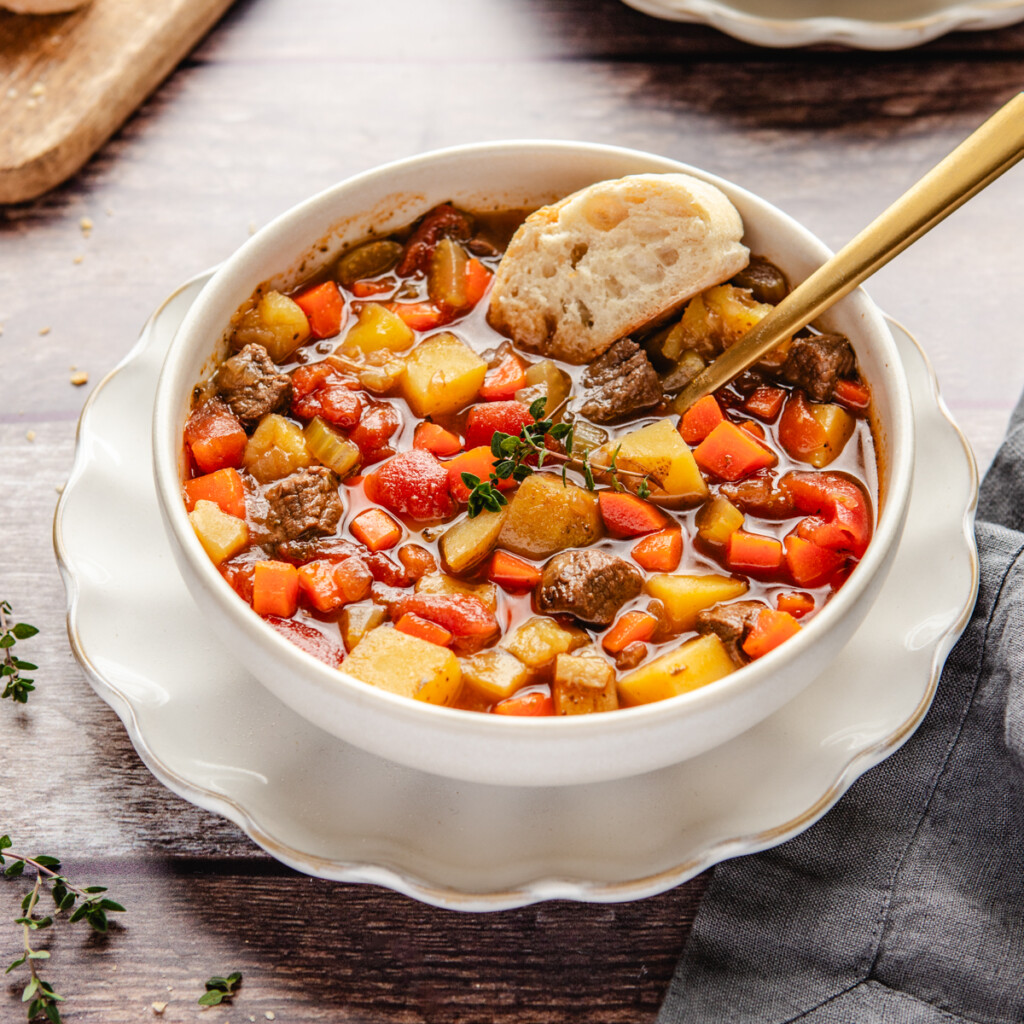 Venison stew in a white bowl surrounded by crusty bread and thyme