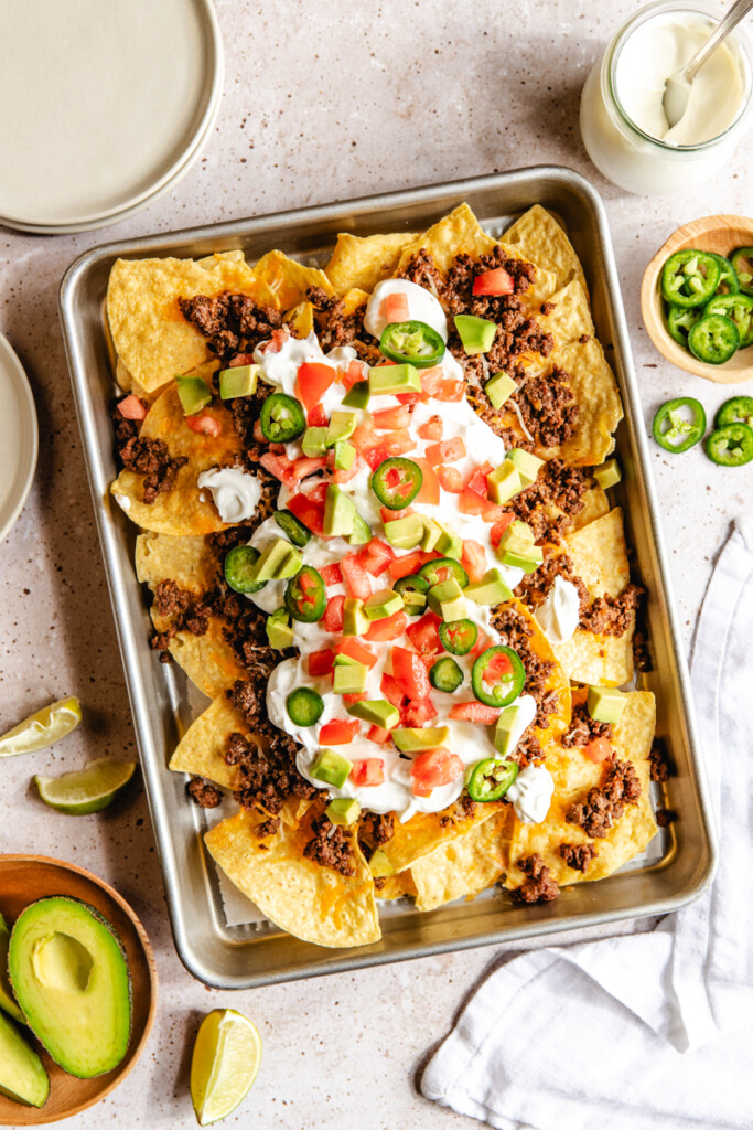 Ground venison taco nachos on a baking sheet with sour cream, tomatoes, jalapeños and avocados