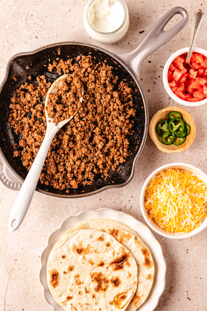 Ground venison meat in a skillet with tortillas, cheese, tomatoes, jalapeños and sour cream in bowls