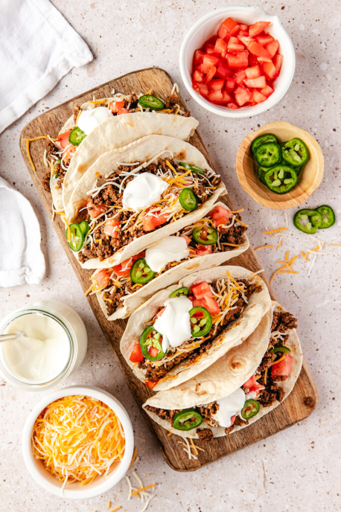 Five ground venison tacos on a wooden cutting board surrounded by bowls of ingredients