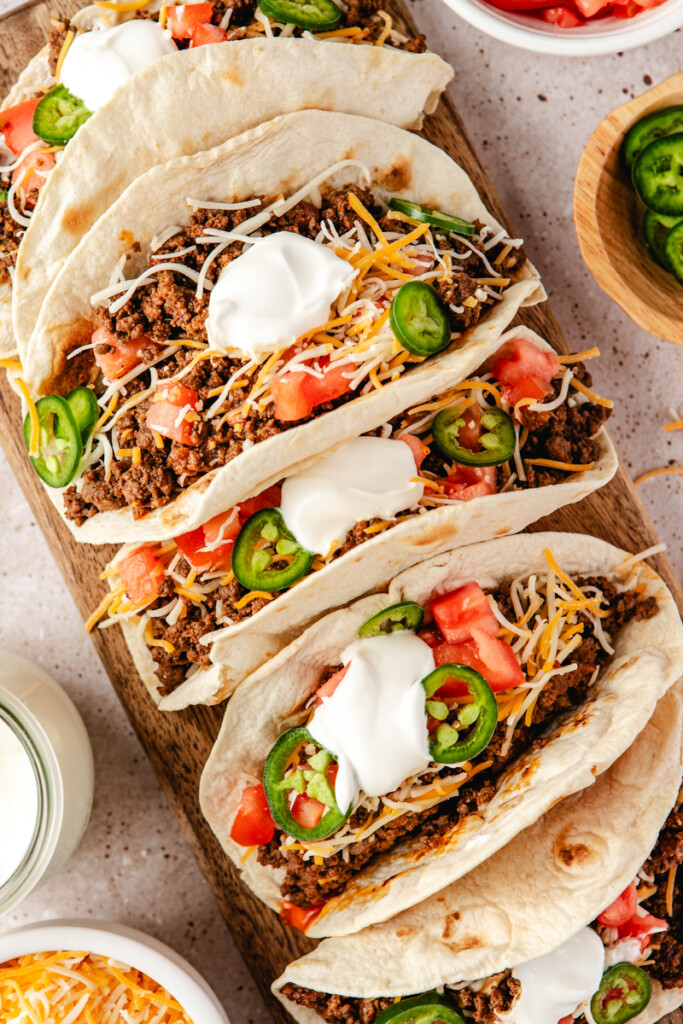 Ground Venison Tacos on a cutting board topped with cheese, tomatoes, jalapeños and sour cream